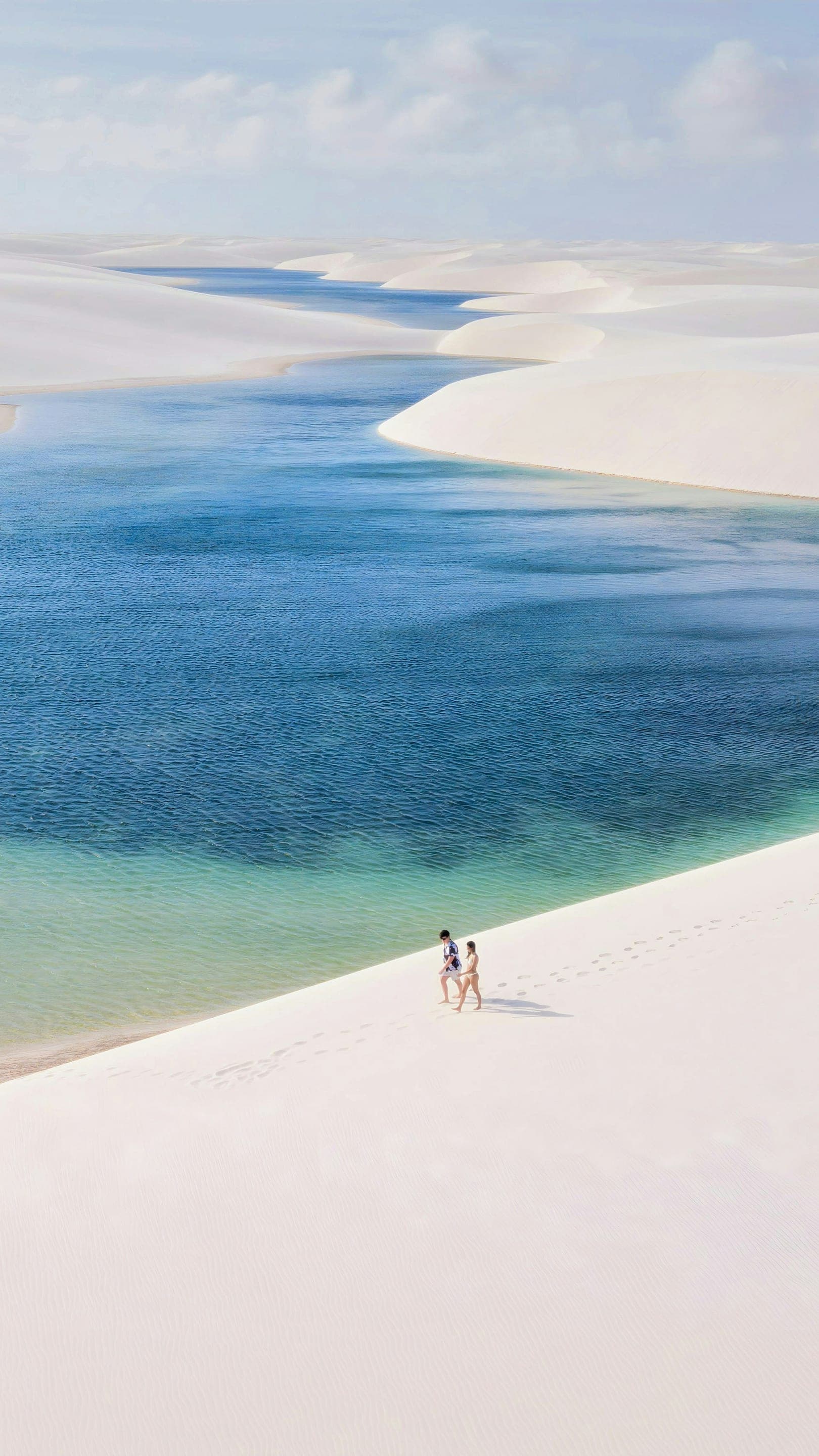Casal caminhando na crista de uma duna branca dos Lençóis Maranhenses, com lagoas azuis e verdes entre as dunas ao redor